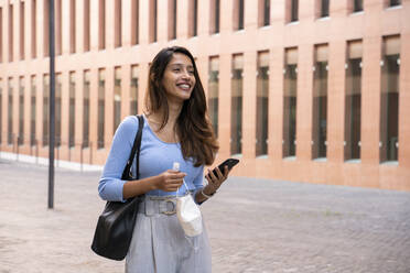 Smiling young businesswoman with smart phone holding protective face mask and hand sanitizer on footpath - AFVF07475