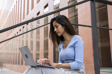 Beautiful businesswoman using laptop while sitting outside office building - AFVF07476