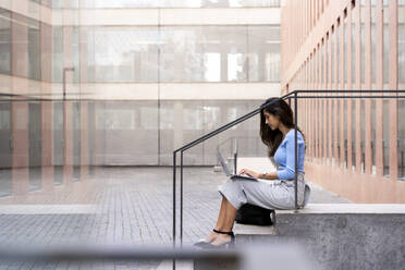 Geschäftsfrau arbeitet an einem Laptop, während sie auf den Stufen vor einem Bürogebäude sitzt - AFVF07479