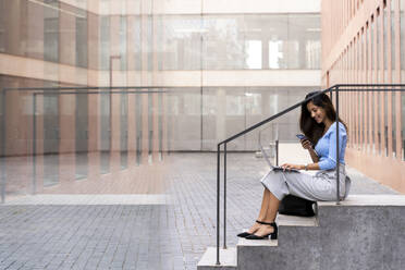 Lächelnde Geschäftsfrau, die ein Smartphone benutzt, während sie mit einem Laptop auf den Stufen vor einem Bürogebäude sitzt - AFVF07481