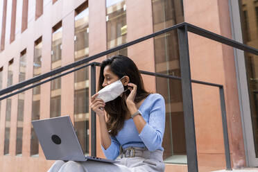 Junge Geschäftsfrau mit Schutzmaske bei der Arbeit mit dem Laptop vor einem Bürogebäude während einer Pandemie - AFVF07488