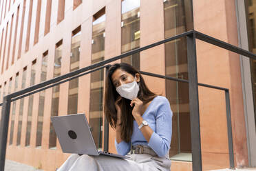 Geschäftsfrau mit Schutzmaske bei der Arbeit mit dem Laptop vor einem Bürogebäude während COVID-19 - AFVF07489