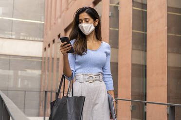 Businesswoman in face mask using smart phone while walking against office building during pandemic - AFVF07490