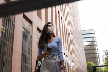 Geschäftsfrau mit Gesichtsmaske schaut weg, während sie an einem Bürogebäude vorbeigeht, während COVID-19 - AFVF07492