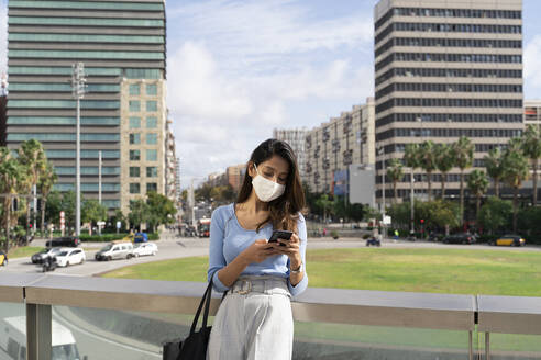 Businesswoman in protective face mask using smart phone while leaning on railing during coronavirus outbreak - AFVF07493