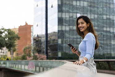 Glückliche junge Geschäftsfrau mit Mobiltelefon auf einer Fußgängerbrücke - AFVF07500