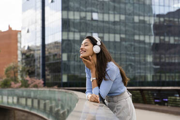 Smiling businesswoman listening music through headphones while leaning on railing of footbridge - AFVF07505