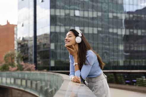 Lächelnde Geschäftsfrau, die Musik über Kopfhörer hört, während sie sich an das Geländer einer Fußgängerbrücke lehnt - AFVF07505