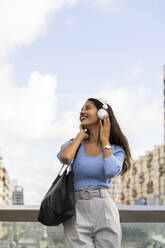 Glückliche Geschäftsfrau, die Musik über Kopfhörer hört, während sie sich an das Geländer gegen den Himmel lehnt - AFVF07508