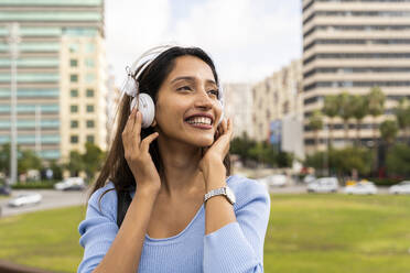 Cheerful young businesswoman listening music through headphones against city - AFVF07511