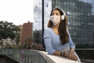 Businesswoman in protective face mask listening music on headphones while leaning on railing - AFVF07512