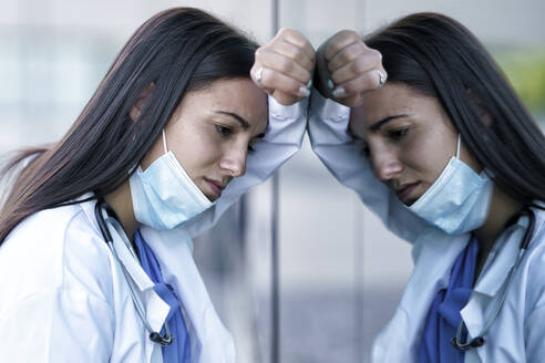 Tired doctor wearing face mask leaning on glass wall of hospital - GGGF00038