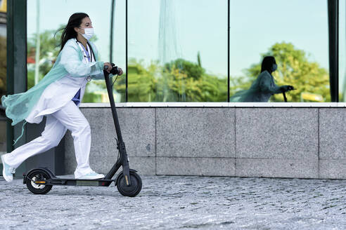 Young doctor in face mask and uniform riding push scooter while going to hospital during COVID-19 - GGGF00040