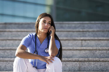 Junger Arzt, der mit seinem Handy telefoniert, während er auf der Treppe vor dem Krankenhaus sitzt - GGGF00042