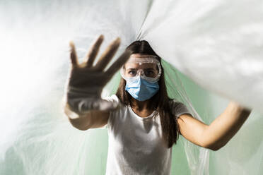 Young woman wearing protective face mask doing stop gesture while covered in plastic during COVID-19 - GIOF09523