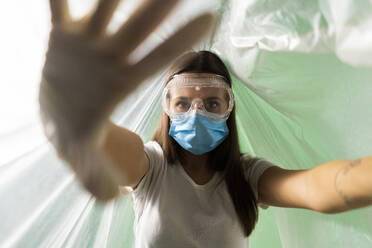 Woman wearing protective face mask doing stop gesture while covered in plastic during coronavirus - GIOF09524