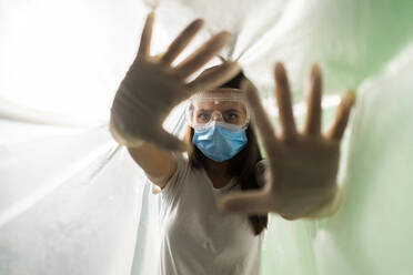 Woman wearing protective face mask doing stop gesture while covered in plastic during COVID-19 outbreak - GIOF09525