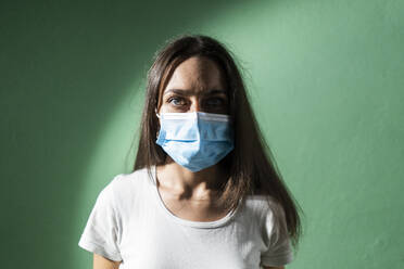 Young woman wearing protective face mask against green wall during COVID-19 - GIOF09526