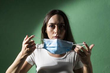 Young woman holding protective face mask against green wall during coronavirus outbreak - GIOF09527