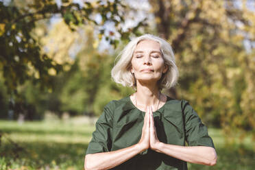 Reife Frau sitzt mit verschränkten Händen beim Yoga im Park - VYF00240
