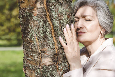 Frau mit geschlossenen Augen umarmt Baum, während sie im Park steht, lizenzfreies Stockfoto