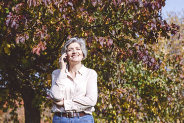 Lächelnde reife Frau, die mit ihrem Handy telefoniert, während sie im Park an einem Baum steht - VYF00246