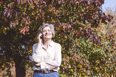 Smiling mature woman talking on mobile phone while standing against tree at park - VYF00246