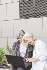 Lächelnde Geschäftsfrauen, die an einem Laptop arbeiten, während sie an einem Gebäude sitzen - VYF00249