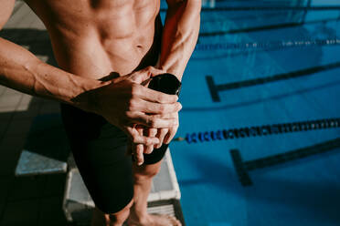 Male swimmer checking smart watch before while standing at poolside - EBBF01284