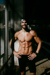 Smiling male swimmer standing by window in locker room - EBBF01303