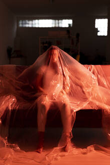 Woman covered in plastic sitting on sofa with fear of coronavirus - GIOF09536