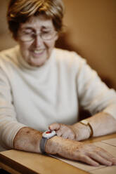 Senior woman pressing emergency button on wrist over table at home - JATF01258