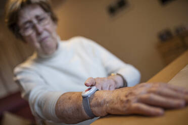 Wrinkled worried woman pressing emergency push button on wrist over table at home - JATF01261