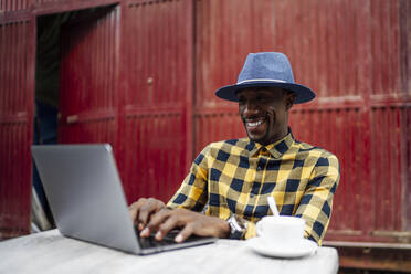 Lächelnder Mann mit Laptop am Tisch vor roter Metallstruktur - PGF00163