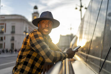Lächelnder afrikanischer Mann mit Smartphone, der wegschaut, während er bei Sonnenuntergang in der Stadt steht - PGF00167