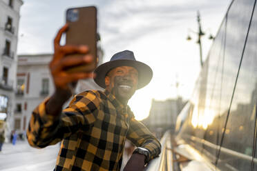 Lächelnder afrikanischer Mann, der mit seinem Smartphone fotografiert, während er bei Sonnenuntergang in der Stadt steht - PGF00168