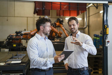 Businessmen with digital tablet discussing while standing against robotic arm machine at factory - DIGF12941