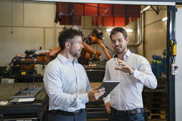 Geschäftsleute mit digitalem Tablet diskutieren, während sie in einer Fabrik vor einer Roboterarm-Maschine stehen - DIGF12941