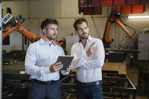 Geschäftsmann zeigt auf einen Kollegen, während er in einer Fabrik gegen eine automatisierte Roboterarm-Maschine steht - DIGF12942