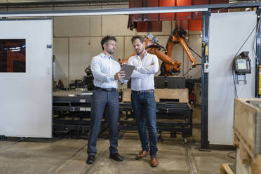 Geschäftsleute, die ein digitales Tablet benutzen, während sie in einer Fabrik vor einer automatisierten Robotermaschine stehen - DIGF12943