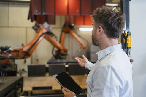 Älterer Geschäftsmann mit digitalem Tablet, der in Richtung einer Roboterarm-Maschine gestikuliert, während er in einer Fabrik steht - DIGF12947