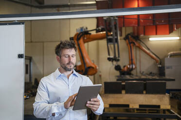 Reifer Geschäftsmann, der ein digitales Tablet benutzt, während er in einer Fabrik vor einer Roboterarm-Maschine steht - DIGF12948
