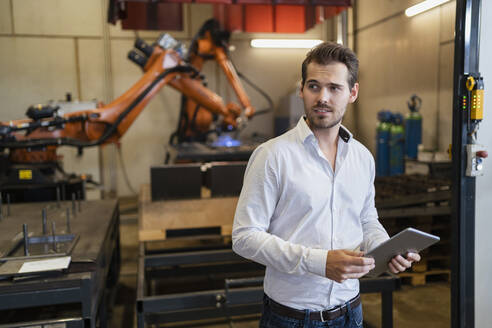 Junger Geschäftsmann, der ein digitales Tablet benutzt, während er vor einer Roboterarm-Maschine in einer modernen Fabrik steht - DIGF12952