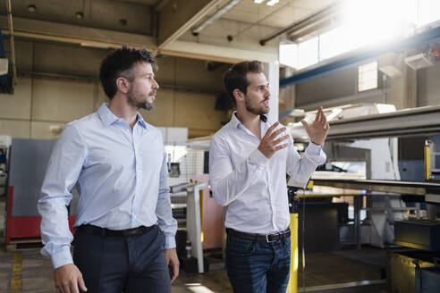 Junger Geschäftsmann schaut weg, während er mit einem Kollegen in einer modernen Fabrik diskutiert - DIGF12959