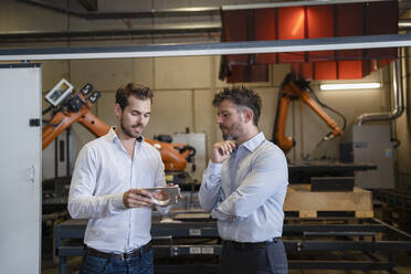 Geschäftsmann mit Metallgegenstand, der mit einem Kollegen diskutiert, während er in einer Fabrik vor einer Robotermaschine steht - DIGF12968