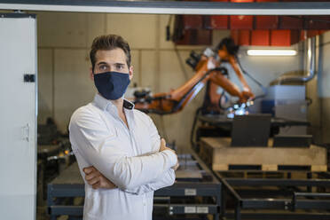 Geschäftsmann mit Gesichtsmaske steht mit verschränkten Armen vor einem Roboterarm in einer Fabrik - DIGF12974