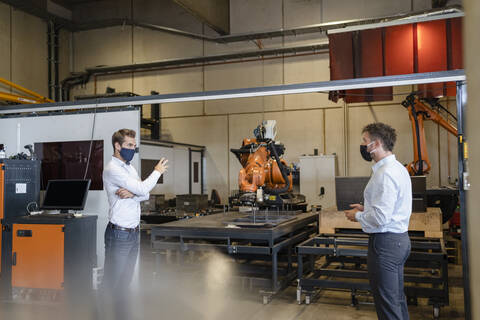 Businessmen with face mask discussing while standing at social distance against robotic arm equipment in factory stock photo