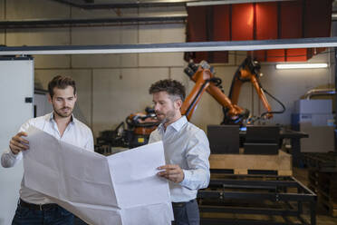 Ein Team mit einer Blaupause diskutiert, während es in einer Fabrik vor einem Roboterarm steht - DIGF12980