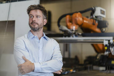 Confident mature businessman standing against automated robotic arm machine at factory - DIGF12988