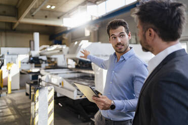 Junger Geschäftsmann mit digitalem Tablet, der in Richtung Ausrüstung gestikuliert, während er neben einem Kollegen in einer Fabrik steht - DIGF12995